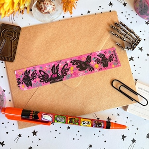 Puede incluir: Una pegatina rectangular de color rosa con un patr&oacute;n repetitivo de ilustraciones de polillas negras con ojos blancos, rodeadas de peque&ntilde;as formas florales y abstractas de colores. La pegatina incluye el texto LYDIAJEANART.
