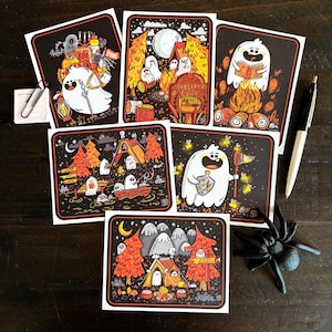 May include: Five rectangular art prints featuring cartoon ghosts in various camping scenes with orange, yellow, and black colour palettes. The illustrations include ghosts by a campfire, in a boat, and near tents. A black pen and a plastic spider are nearby.