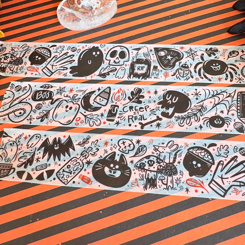May include: Three rolls of decorative washi tape with Halloween-themed illustrations. The black and white drawings include ghosts, bats, skulls, spiders, and the text "Creep It Real". The tape has a light blue background and is on an orange and black striped surface.