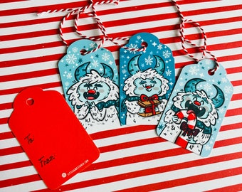 May include: Three blue and white gift tags with a wintery theme. Each tag features a cartoon yeti with a different expression. The tags are decorated with snowflakes and have the words "To:" and "From:" printed on them.