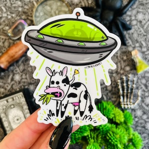 May include: A black and white cow with pink udders is being abducted by a flying saucer with a green interior. The saucer is emitting green beams of light. The cow is eating grass.