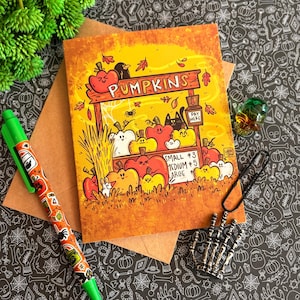 May include: A rectangular greeting card featuring a colorful illustration of a pumpkin patch stand with the text PUMPKINS, PAY HERE, SMALL $3, MEDIUM $5, and LARGE. The card is accompanied by a bright green pen with pumpkin motifs and a skeleton hand clip.