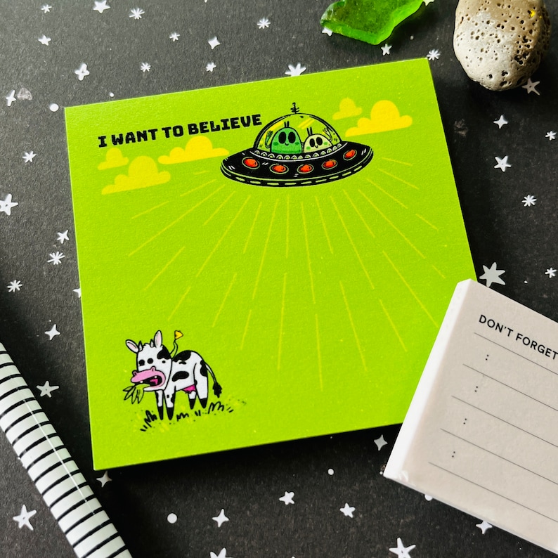 May include: Green notepad with a black and white cow illustration and a cartoon UFO with green aliens. The notepad has the text "I WANT TO BELIEVE" at the top and a smaller notepad with the text "DON'T FORGET" on the right side.