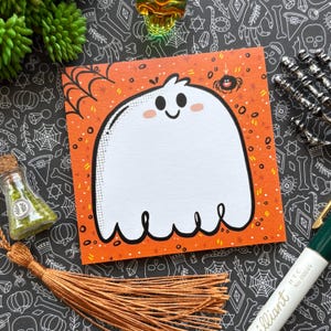 May include: Square orange sticky note with a cartoon ghost illustration for Halloween. The ghost has a smiling face and is surrounded by a spider web and a small spider. The background is decorated with Halloween patterns.