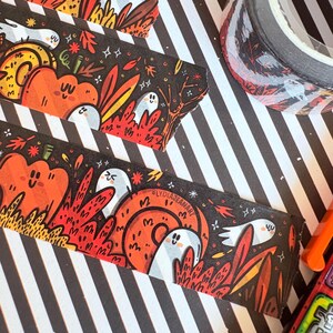 Pu&ograve; includere: Nastro adesivo decorativo a tema Halloween. Il nastro presenta zucche arancioni, fantasmi bianchi e fogliame rosso su sfondo nero. Un rotolo di nastro &egrave; visibile su una superficie a strisce bianche e nere.