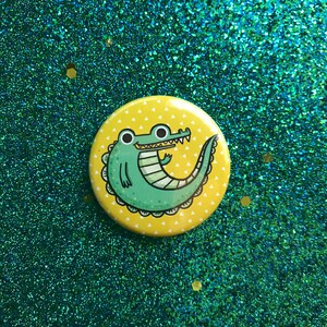 May include: A yellow button with a green cartoon alligator with white teeth and a white triangle pattern on a yellow background.