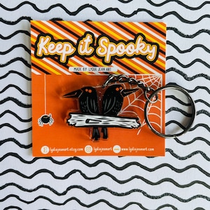 May include: A black and white acrylic keychain featuring two crows perched on a branch. The keychain has a silver metal ring and the text "Keep it Spooky" on a yellow and orange striped background. The text "Made by: Lydia Jean Art" is also included.