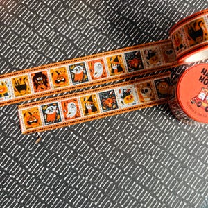 Puede incluir: Un rollo de cinta decorativa naranja y blanca con un patr&oacute;n repetitivo de personajes de Halloween que incluye un gato negro, calabaza, vampiro, fantasma, bruja, ara&ntilde;a y cuervo, con una etiqueta redonda naranja que dice Happy Halloween.