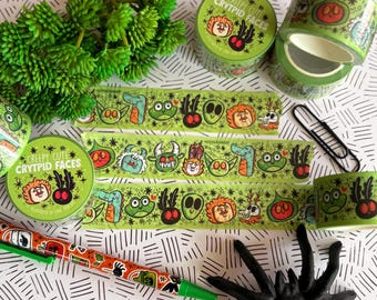 Puede incluir: Una colección de rollos de cinta decorativa verde y un bolígrafo con un patrón repetitivo de caras de monstruos de dibujos animados. El empaque muestra el texto "CREEPY CUTE CRYPTID FACES" junto a varias cabezas de criaturas ilustradas.