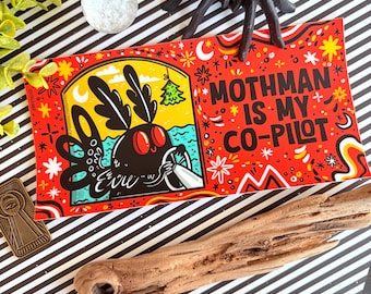 May include: A red sticker with the text "MOTHMAN IS MY CO-PILOT" and a cartoon Mothman illustration. The sticker has a black and white striped background with a decorative spider, a white stone, a wooden stick, and a gold-colored pin.