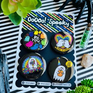 May include: A set of four round, black enamel pins with colorful ghost and rainbow designs. One pin says "Be You", another says "Love is Love", and a third says "Less Hate More Spooks". The pins are on a black and white scalloped card.