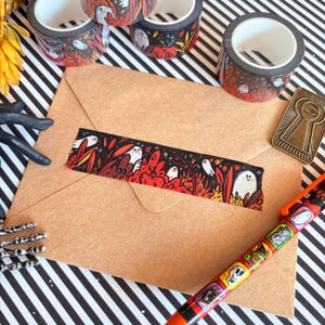 Pu&ograve; includere: Un assortimento di articoli di cancelleria a tema Halloween. Diversi rotoli di nastro adesivo decorativo con sfondo nero e un motivo ripetuto di fantasmi bianchi e fogliame rosso. Una busta marrone con una striscia di nastro abbinato, una penna e un ciondolo a forma di buco della serratura dorato.
