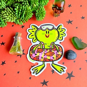 May include: A quirky sticker featuring a lime green cartoon frog wearing swim goggles and a patterned inner tube. The sticker is surrounded by decorative elements on a red background with black stars.