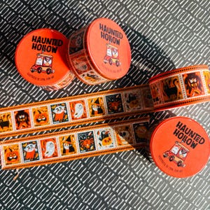 Puede incluir: Tres latas redondas de color naranja etiquetadas como Haunted Hollow con una ilustraci&oacute;n de veh&iacute;culo, acompa&ntilde;adas de dos tiras de cinta decorativa con paneles cuadrados repetidos de personajes de Halloween como fantasmas, calabazas y gatos.