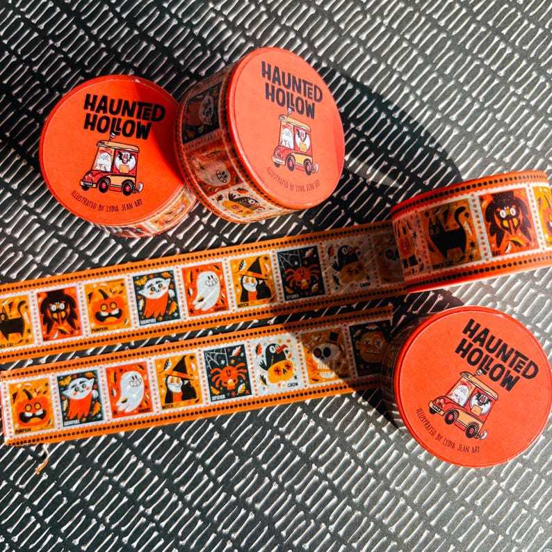 Puede incluir: Tres latas redondas de color naranja etiquetadas como Haunted Hollow con una ilustraci&oacute;n de veh&iacute;culo, acompa&ntilde;adas de dos tiras de cinta decorativa con paneles cuadrados repetidos de personajes de Halloween como fantasmas, calabazas y gatos.