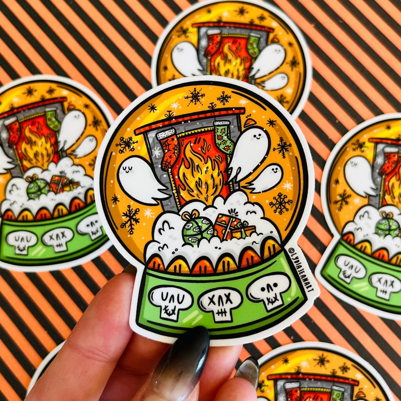 May include: A round, colorful sticker with a snow globe design. The snow globe contains a fireplace with stockings, a fire, and ghosts. The base of the snow globe is green and has the words "UUU" and "XXX" on it.