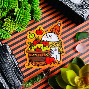 Puede incluir: Una pegatina con un lindo fantasma de dibujos animados que lleva un sombrero rojo y blanco y una bufanda. El fantasma sostiene una cesta llena de manzanas rojas y verdes. La pegatina tiene un tema otoñal.