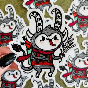 May include: A gray and red cartoon monster with horns, a red scarf, and a backpack. The monster is holding a branch in its right hand and has a tongue sticking out. The sticker is white with a black outline.