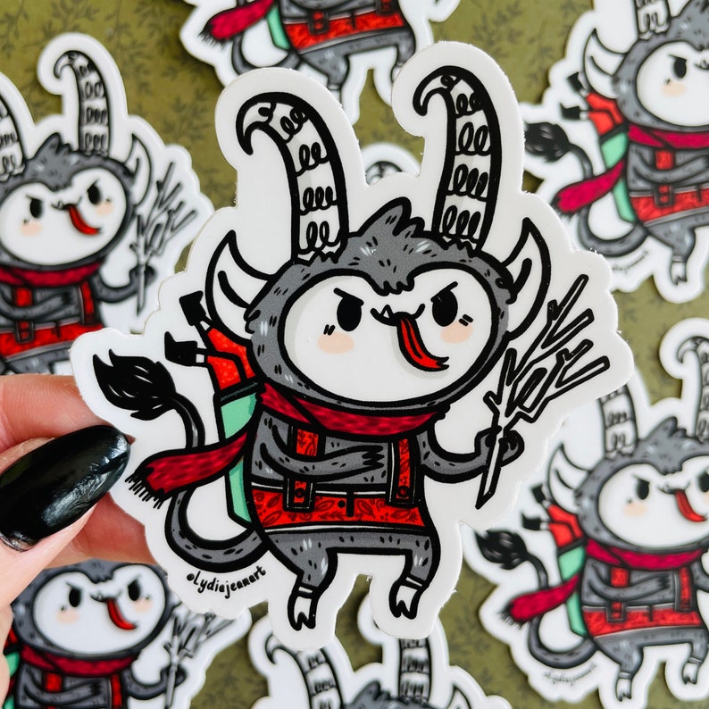May include: A gray and red cartoon monster with horns, a red scarf, and a backpack. The monster is holding a branch in its right hand and has a tongue sticking out. The sticker is white with a black outline.