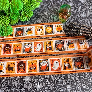 Puede incluir: Un rollo de cinta decorativa washi con un patr&oacute;n repetitivo de cuadrados naranjas y negros con tem&aacute;tica de Halloween. Cada cuadrado muestra un personaje o icono &uacute;nico, incluyendo un fantasma, bruja, vampiro, calabaza, ara&ntilde;a y una calavera.