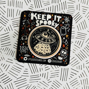 May include: Square black sticker with the words "KEEP IT SPOOKY" in white. The sticker features a UFO with aliens, stars, ghosts, and a skull. The design is in orange and white.