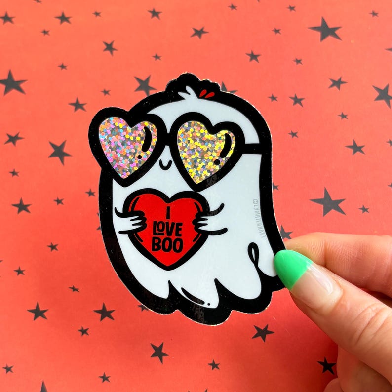 May include: A white ghost sticker with black outlines, wearing heart-shaped sunglasses with glitter. The ghost is holding a red heart with the words "I LOVE BOO" in white. The background is orange with black star shapes.