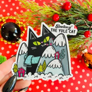 May include: A black cat with green eyes wearing a blue scarf stands in a snowy mountain scene. The cat is holding a red and white candy cane in its mouth. The text "Beware of the Yule Cat" is written in black on a white background.