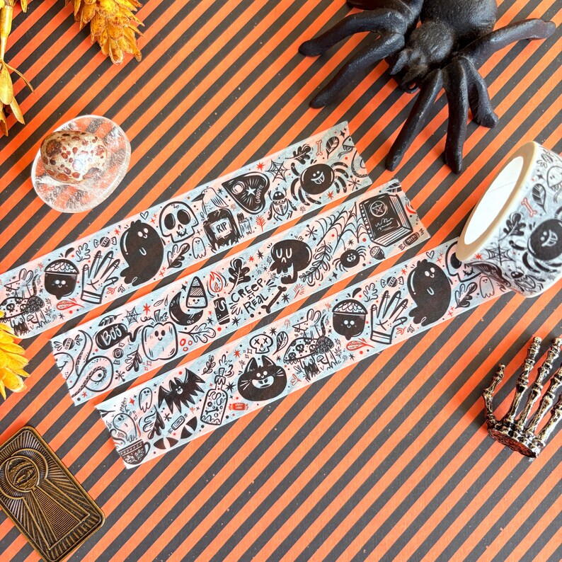 May include: Halloween-themed decorative washi tape with black and white illustrations of ghosts, spiders, and skulls. The tape features the text "Creep Real" and "Boo". The tape is on a striped orange and black background.