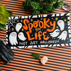Puede incluir: Pegatina en blanco y negro con texto naranja que dice "Spooky for Life Not Just Halloween". La pegatina presenta una telaraña de dibujos animados y varios fantasmas blancos con diferentes expresiones faciales.