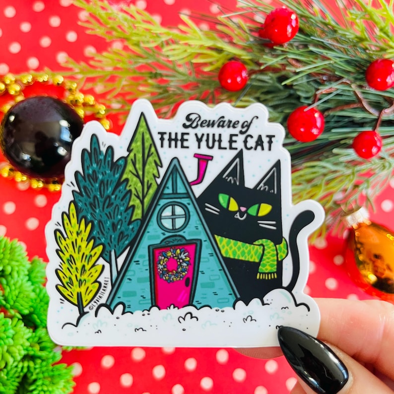 May include: A white sticker with a black cat wearing a green scarf in front of a blue house with a pink door. The text on the sticker reads "Beware of the Yule Cat".