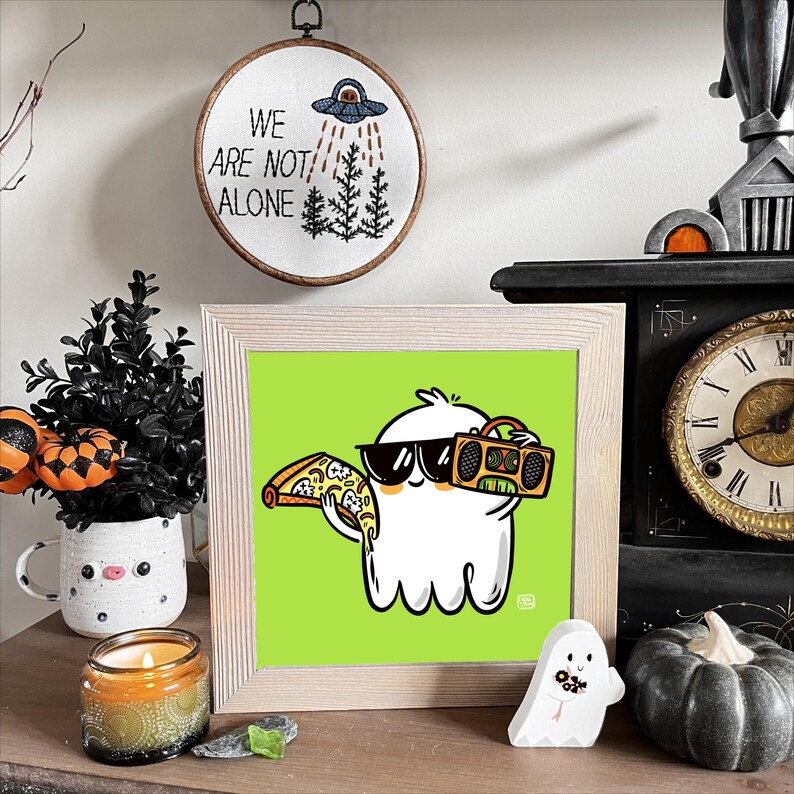 May include: A framed illustration of a white ghost wearing sunglasses and holding a slice of pizza and a boombox. The background is green. The frame is light wood.