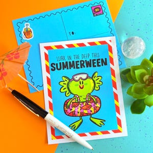 May include: A greeting card with the text "Lurk in the Deep This Summerween" featuring a cartoon frog in a swim ring. A blue postcard and envelope are behind the card, along with a pen and a cocktail umbrella.