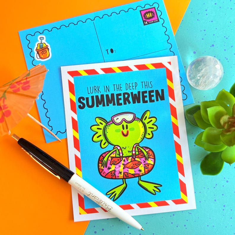 May include: A greeting card with the text "Lurk in the Deep This Summerween" featuring a cartoon frog in a swim ring. A blue postcard and envelope are behind the card, along with a pen and a cocktail umbrella.