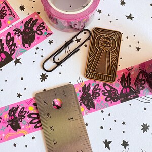 Puede incluir: Un rollo de cinta washi rosa con ilustraciones de polillas negras, un clip de metal negro y un pin esmaltado rectangular dorado que representa un ojo dentro de una cerradura. Una regla de metal muestra una medida de 2,5 cm de ancho.