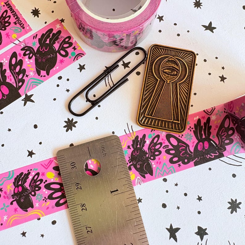 Puede incluir: Un rollo de cinta washi rosa con ilustraciones de polillas negras, un clip de metal negro y un pin esmaltado rectangular dorado que representa un ojo dentro de una cerradura. Una regla de metal muestra una medida de 2,5 cm de ancho.