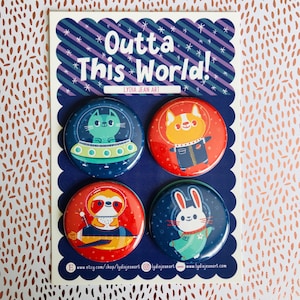 May include: Set of four 1.5 inch diameter round pinback buttons featuring colorful illustrations of a cat in a spaceship, a bunny in a spacesuit, a fox in a spacesuit, and a sloth in a spaceship. The buttons have a white background with black outlines and the text "Outta This World!" and "Lydia Jean Art" printed on the card.