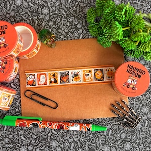 Puede incluir: Una colecci&oacute;n de art&iacute;culos de papeler&iacute;a con tem&aacute;tica Haunted Hollow que incluye varios rollos de cinta decorativa naranja, un sobre marr&oacute;n, un clip negro, un bol&iacute;grafo verde con motivos de Halloween y una peque&ntilde;a figura de mano esquel&eacute;tica.