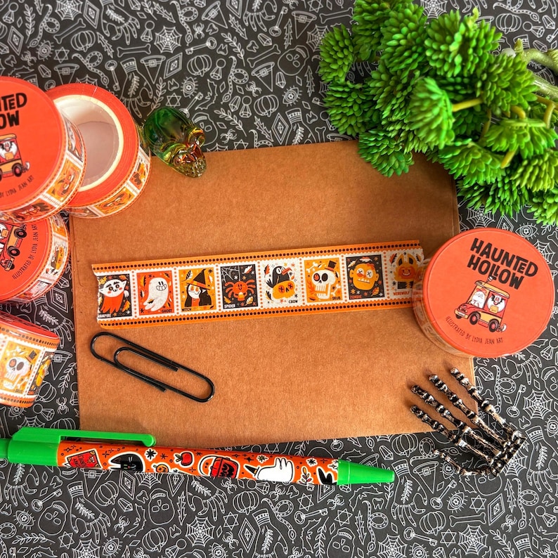 Puede incluir: Una colecci&oacute;n de art&iacute;culos de papeler&iacute;a con tem&aacute;tica Haunted Hollow que incluye varios rollos de cinta decorativa naranja, un sobre marr&oacute;n, un clip negro, un bol&iacute;grafo verde con motivos de Halloween y una peque&ntilde;a figura de mano esquel&eacute;tica.
