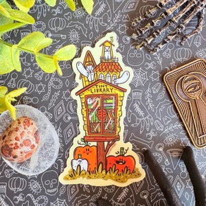 Puede incluir: Una pegatina caprichosa con una pequeña biblioteca amarilla y naranja. La pegatina incluye dos fantasmas, calabazas y el texto "TINY LIBRARY". Ideal para Halloween o amantes de los libros.