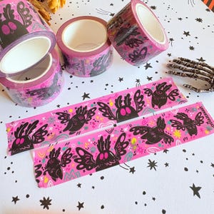 Puede incluir: Tres rollos de cinta washi de color rosa brillante con un patr&oacute;n repetitivo de criaturas aladas negras con ojos grandes. Dos tiras de cinta est&aacute;n extendidas, mostrando el mismo dise&ntilde;o gr&aacute;fico con peque&ntilde;os acentos coloridos y el texto LYDIAJEANART.