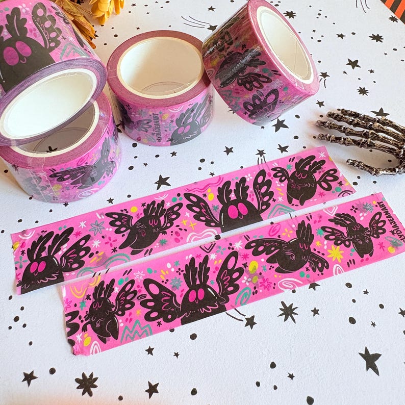 Puede incluir: Tres rollos de cinta washi de color rosa brillante con un patr&oacute;n repetitivo de criaturas aladas negras con ojos grandes. Dos tiras de cinta est&aacute;n extendidas, mostrando el mismo dise&ntilde;o gr&aacute;fico con peque&ntilde;os acentos coloridos y el texto LYDIAJEANART.