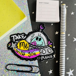 May include: A colorful sticker with the text "Take me with you please" featuring an alien in a spaceship with two cats. A black and white striped pen, a notepad, and a paperclip are also in the image.
