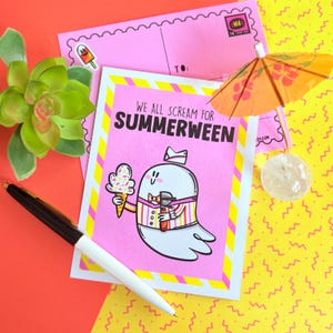 May include: A pink greeting card with the text "WE ALL SCREAM FOR SUMMERWEEN" and a cartoon ghost holding an ice cream cone and a drink. The card is decorated with a yellow and white border and is next to a pink envelope and a pen.