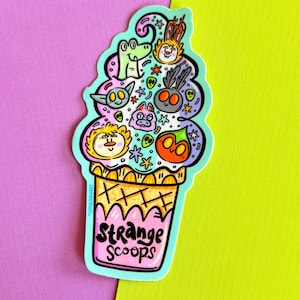 May include: A die-cut sticker shaped like an ice cream cone with a light blue outline. The top features various colorful cartoon creature faces and small stars. The cone is pink with a cross-hatch pattern and the text Strange Scoops.