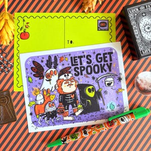 Könnte beinhalten: Eine Halloween-Postkarte mit dem Schriftzug "LET'S GET SPOOKY" und Illustrationen von Cartoon-Monstern. Ein grüner Stift mit Halloween-Designs befindet sich im Vordergrund. Eine limettengrüne Postkarte befindet sich hinter der Hauptkarte.