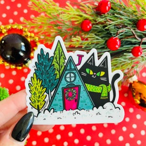 May include: A black cat with green eyes and a green scarf stands in front of a blue and green triangular house with a pink door and a wreath. The house is surrounded by snow and green trees.