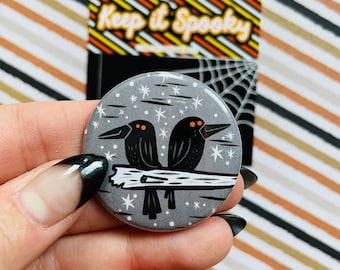 May include: A grey button with a white background featuring two black birds with orange eyes perched on a branch. The background is filled with white stars.