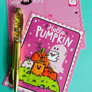 May include: A gold pen with floating stars and a pink postcard that says "Hello Pumpkin" with a cartoon ghost and pumpkins. A second pink postcard is behind the first.