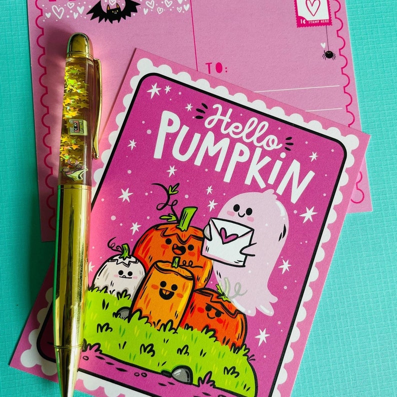 May include: A gold pen with floating stars and a pink postcard that says "Hello Pumpkin" with a cartoon ghost and pumpkins. A second pink postcard is behind the first.