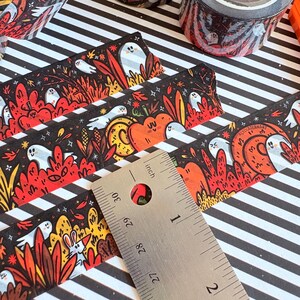 Pu&ograve; includere: Nastro adesivo decorativo a tema Halloween. Il nastro nero presenta illustrazioni di fantasmi, zucche e fogliame in arancione, rosso e giallo. Il nastro &egrave; largo circa 2,5 cm, come mostra un righello. &Egrave; visibile anche un rotolo di nastro.